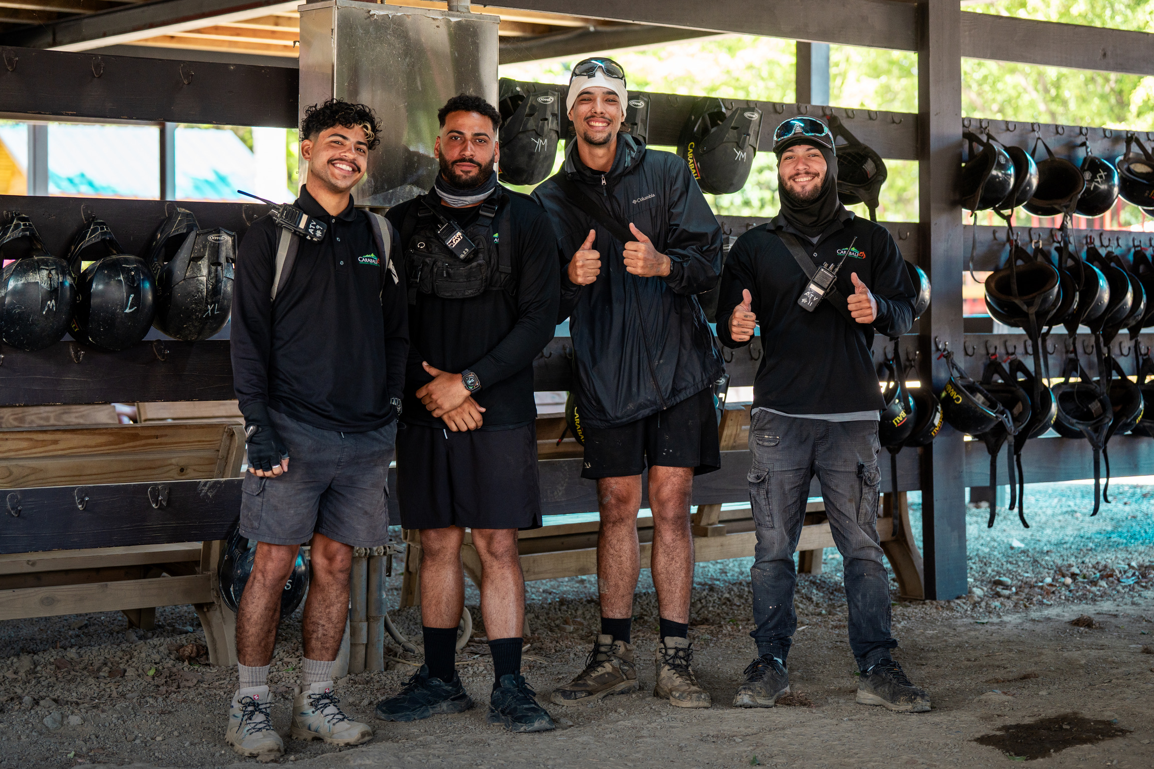 Four smiling men in work attire pose inside a structure with helmets hanging on the walls.