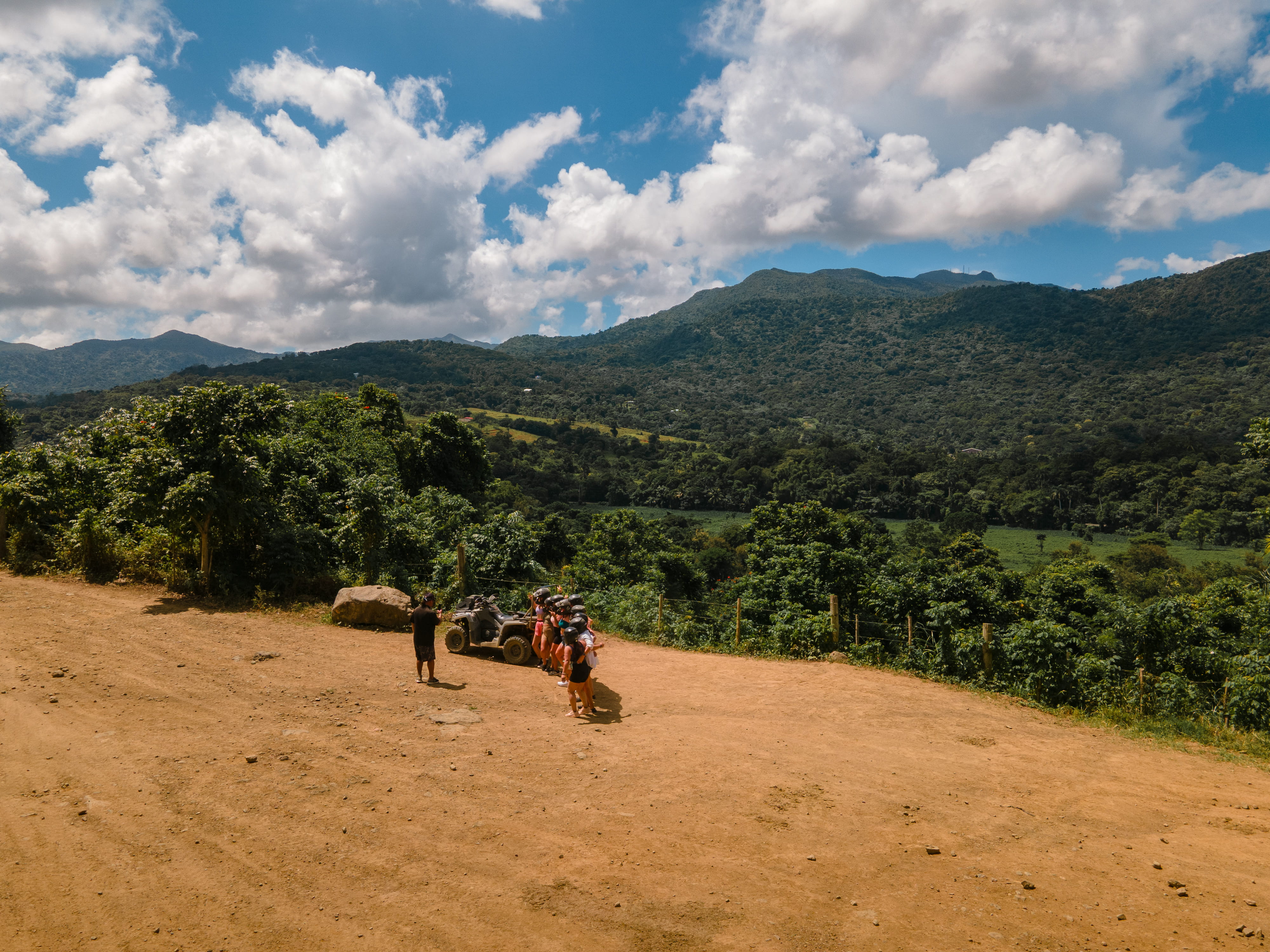 People with ATVs on a dirt road, lush green hills, and cloudy sky in the background.