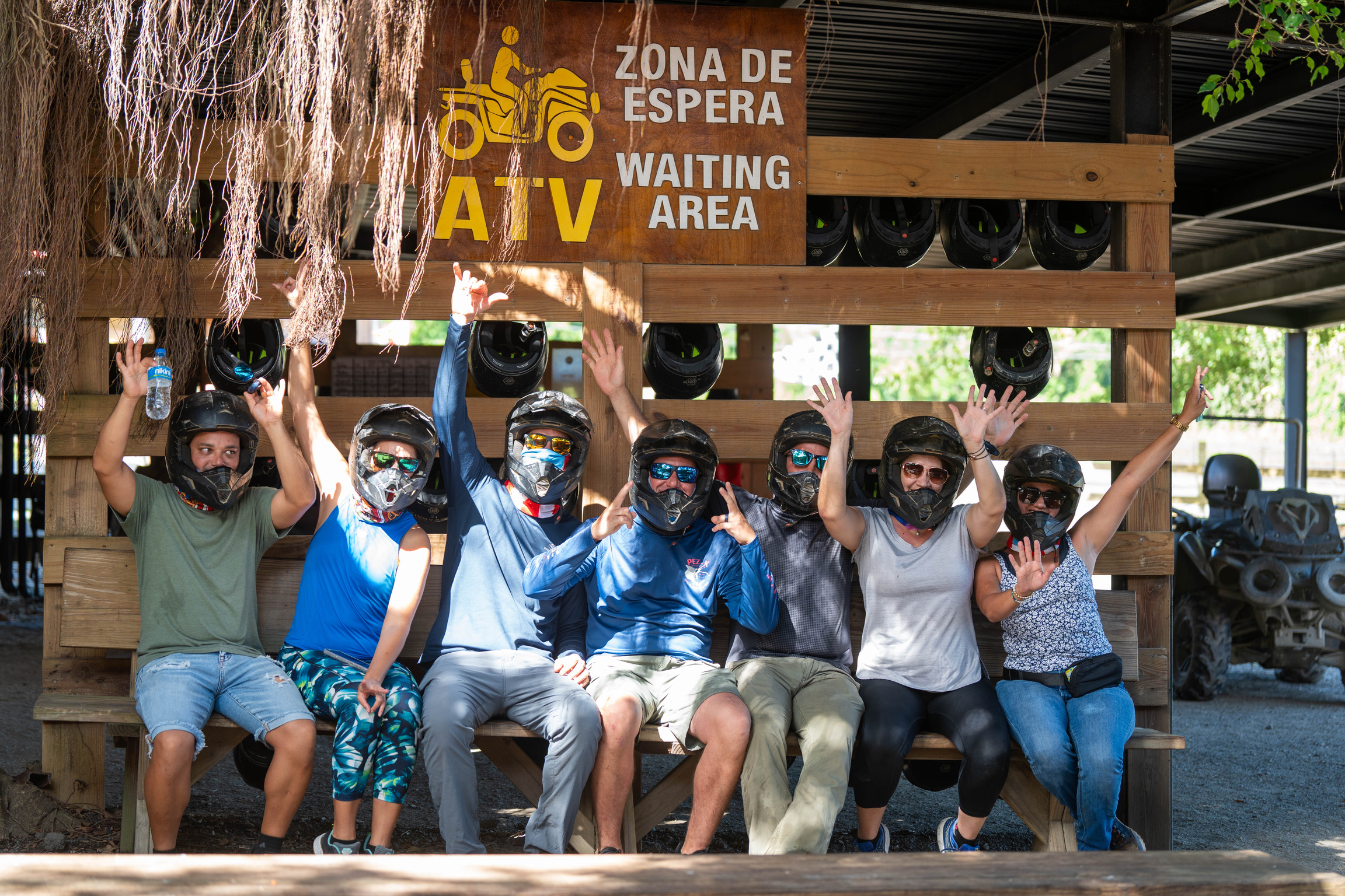 Group sitting with helmets, raising hands, near ATV waiting area sign.