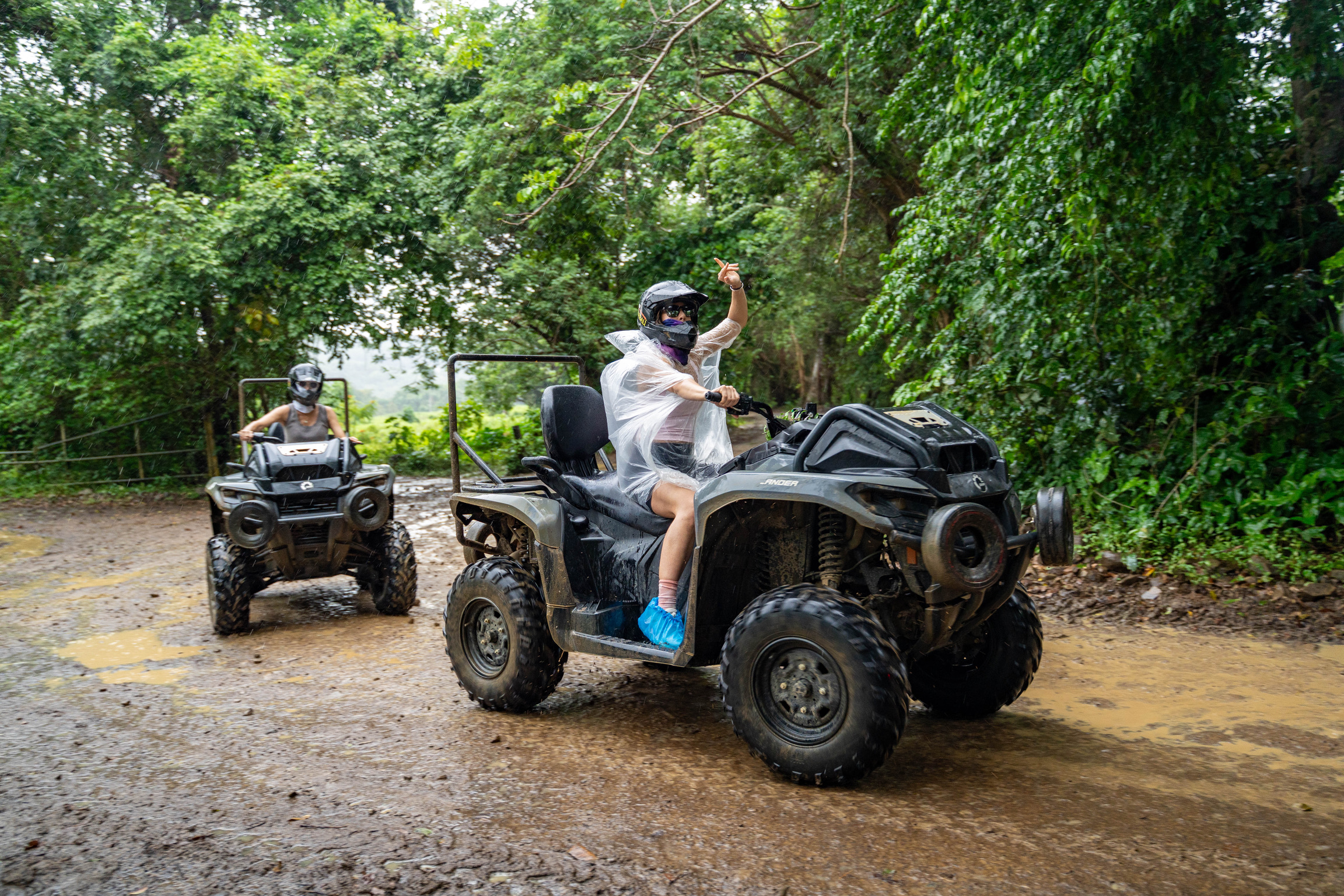 Two people on ATVs riding through a muddy forest trail.