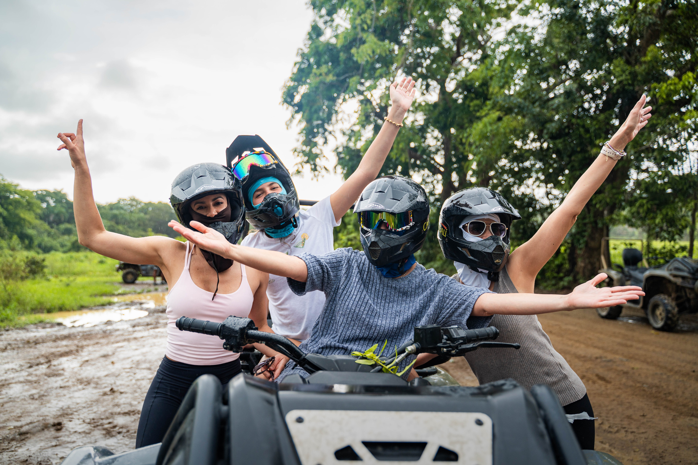 Four people with helmets pose joyfully on an ATV in a lush outdoor setting.