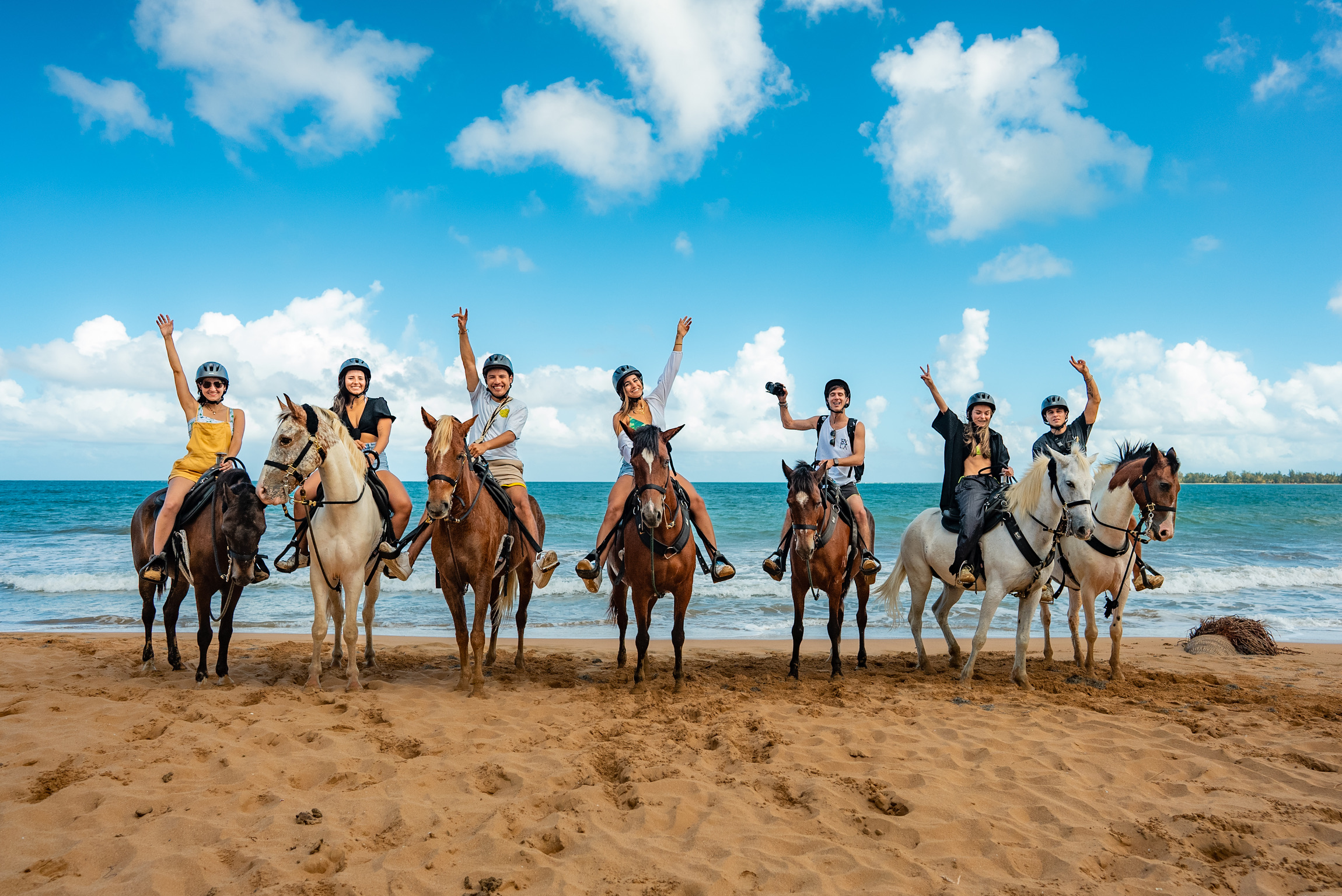 Group horseback riding on beach, hands raised, blue sky.