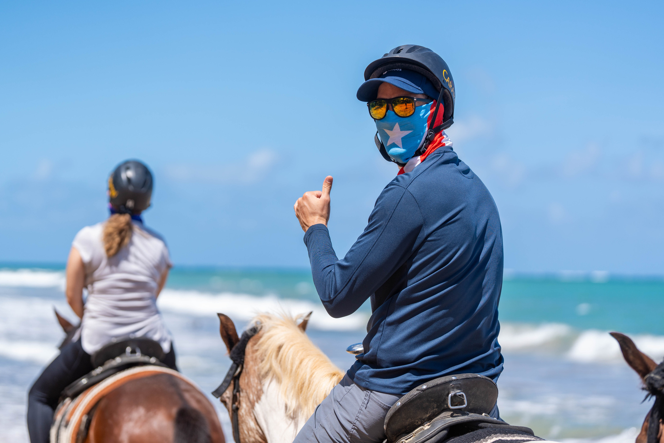 Two people riding horses on a beach, one giving a thumbs up.
