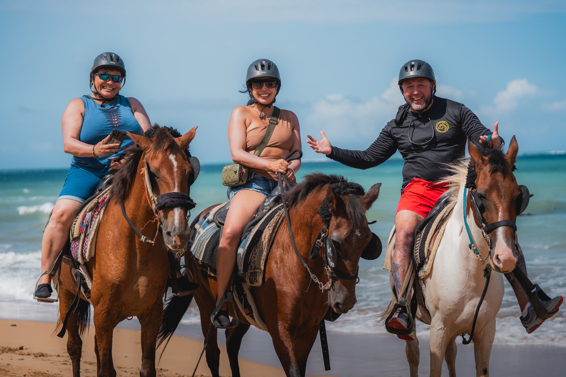 Three people on horseback at the beach, wearing helmets and casual attire.