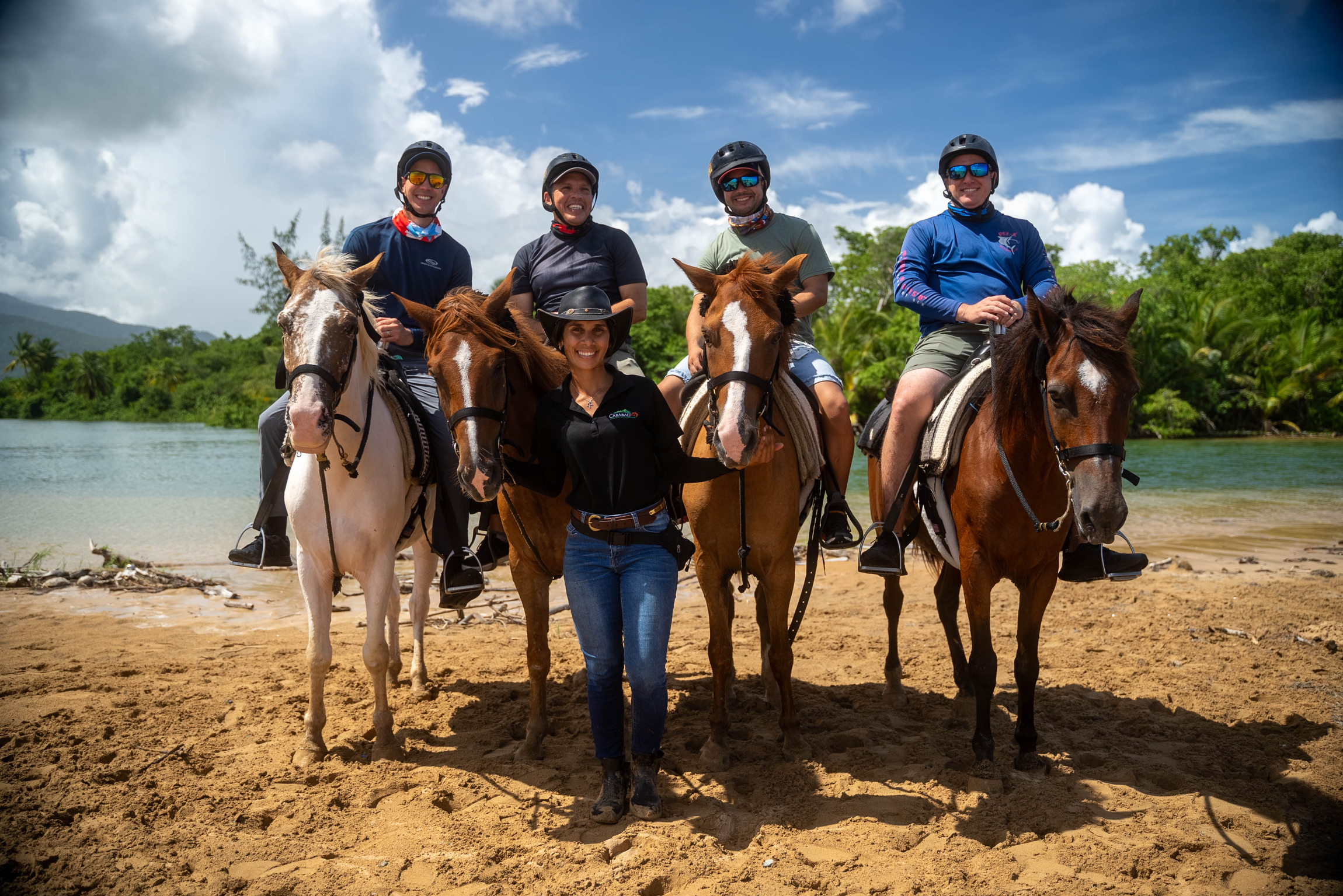 Group of four people on horses and one standing on sandy beach.