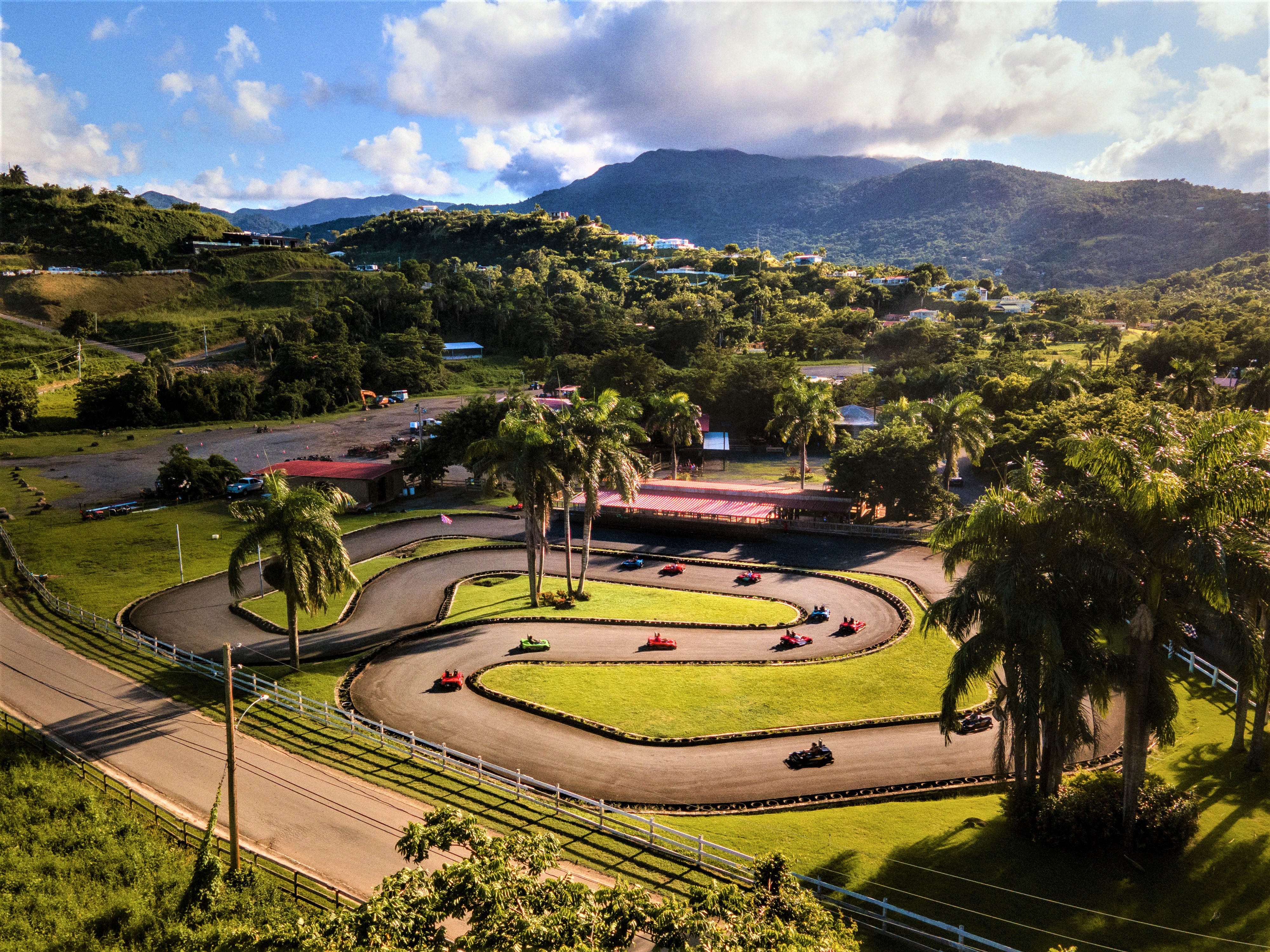 Aerial view of a go-kart track surrounded by trees and hills under a blue sky with clouds.