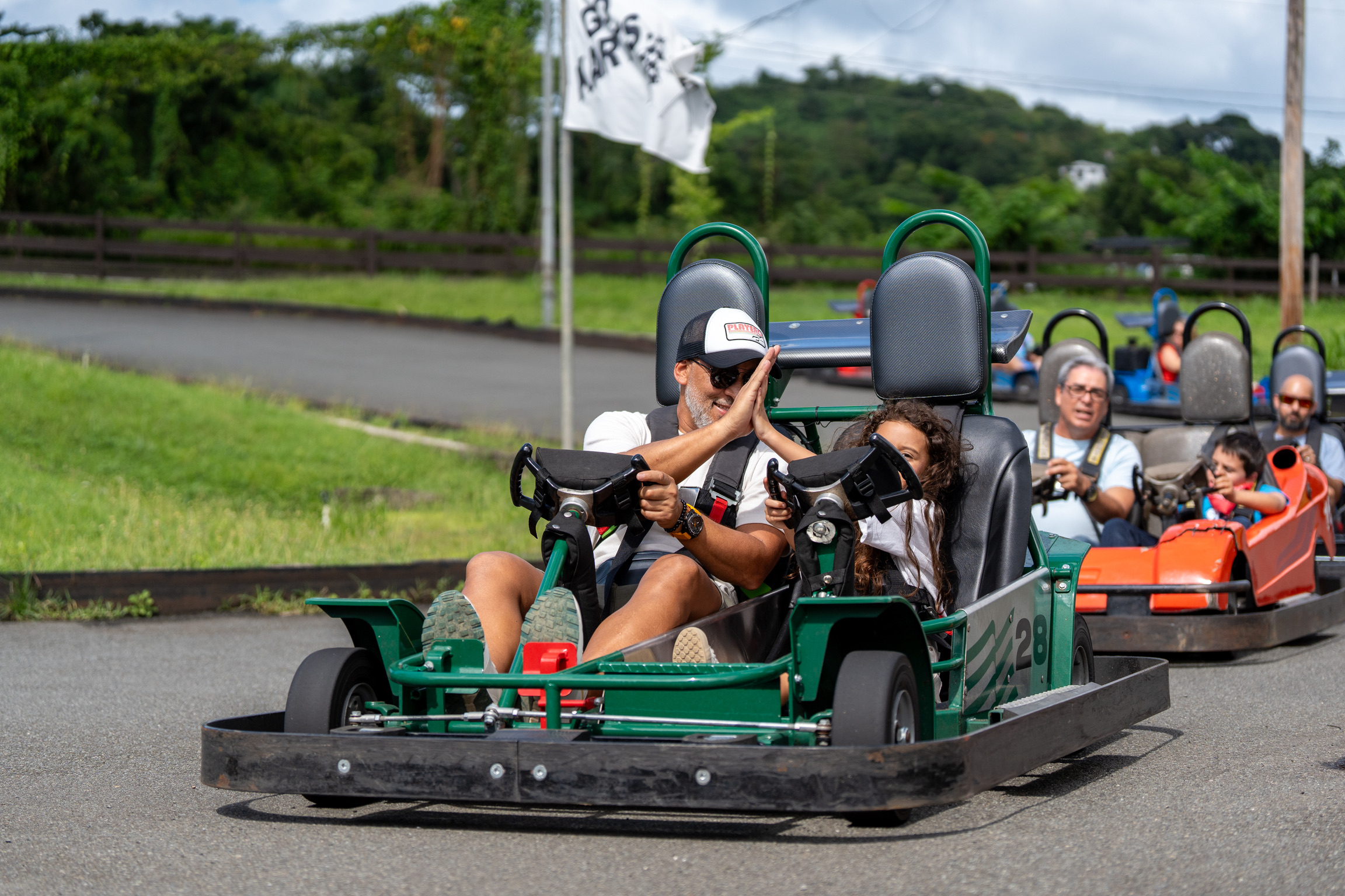 People driving go-karts on an outdoor track with a flag in the background.
