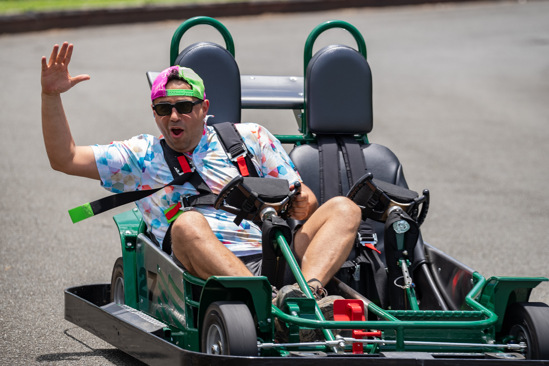 Man in tie-dye shirt waving while driving a green go-kart.