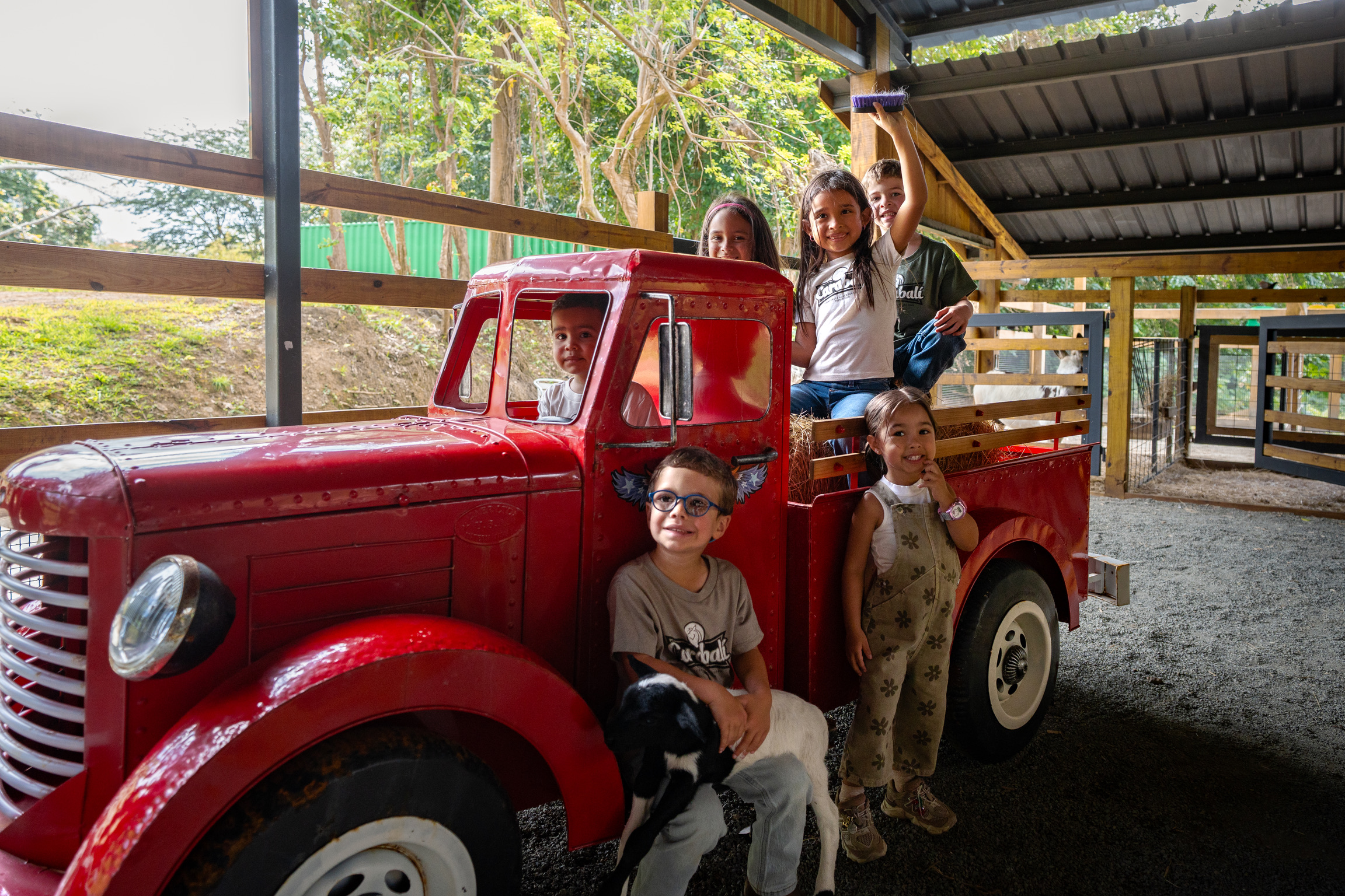 Children smiling and posing with a red vintage truck under a shelter.