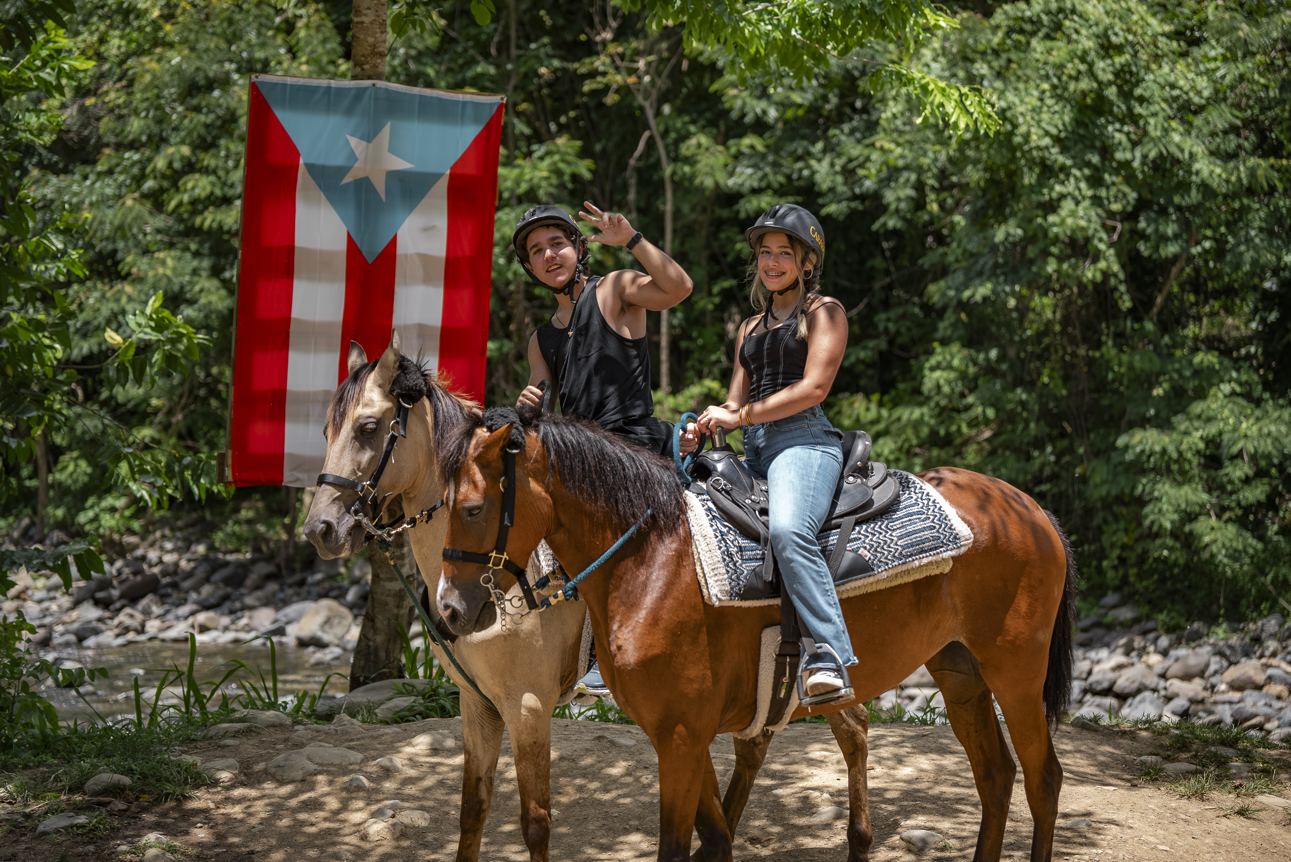 Two people on horses in front of Puerto Rican flag in a forest.