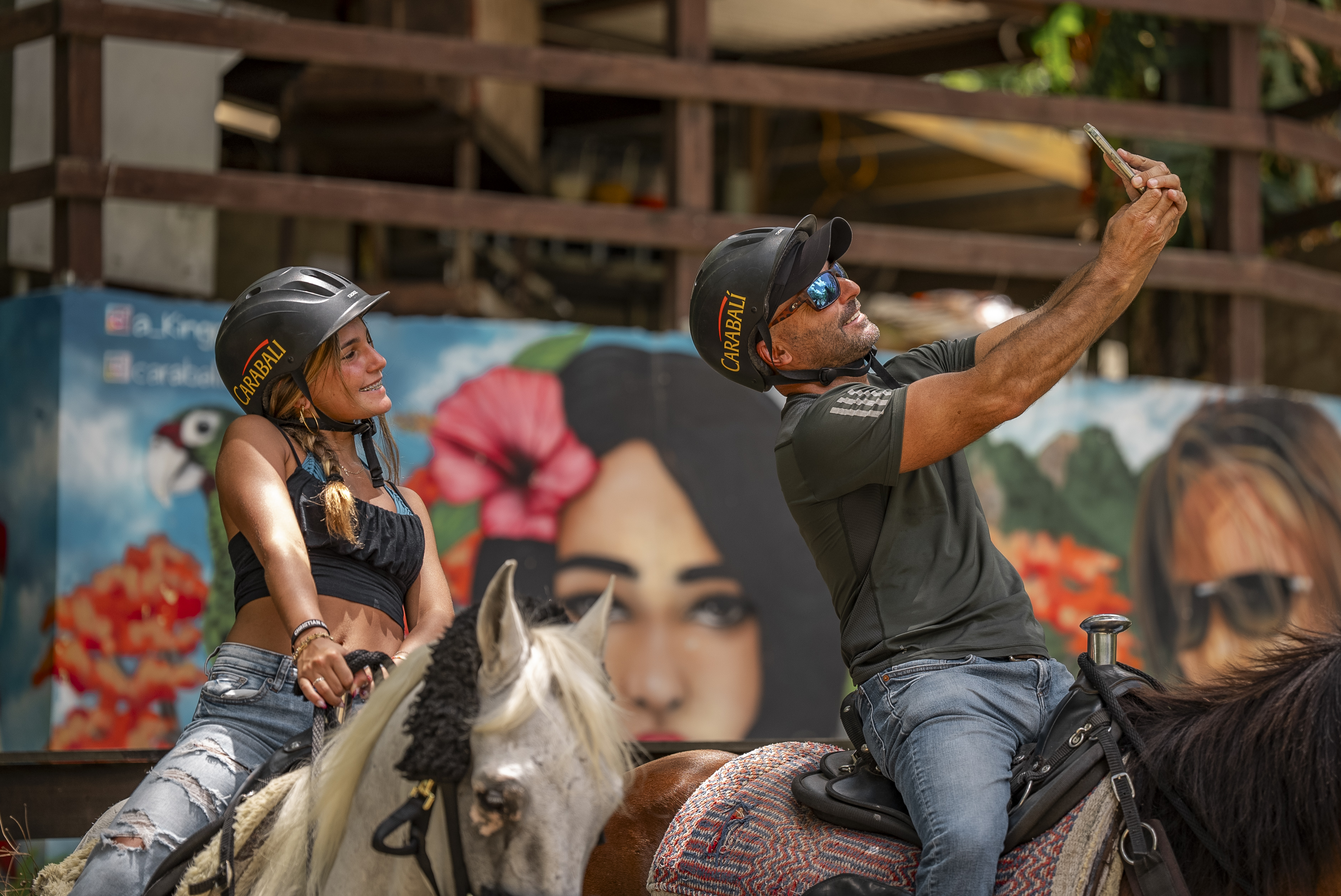 Two people on horses taking a selfie in front of a colorful mural.