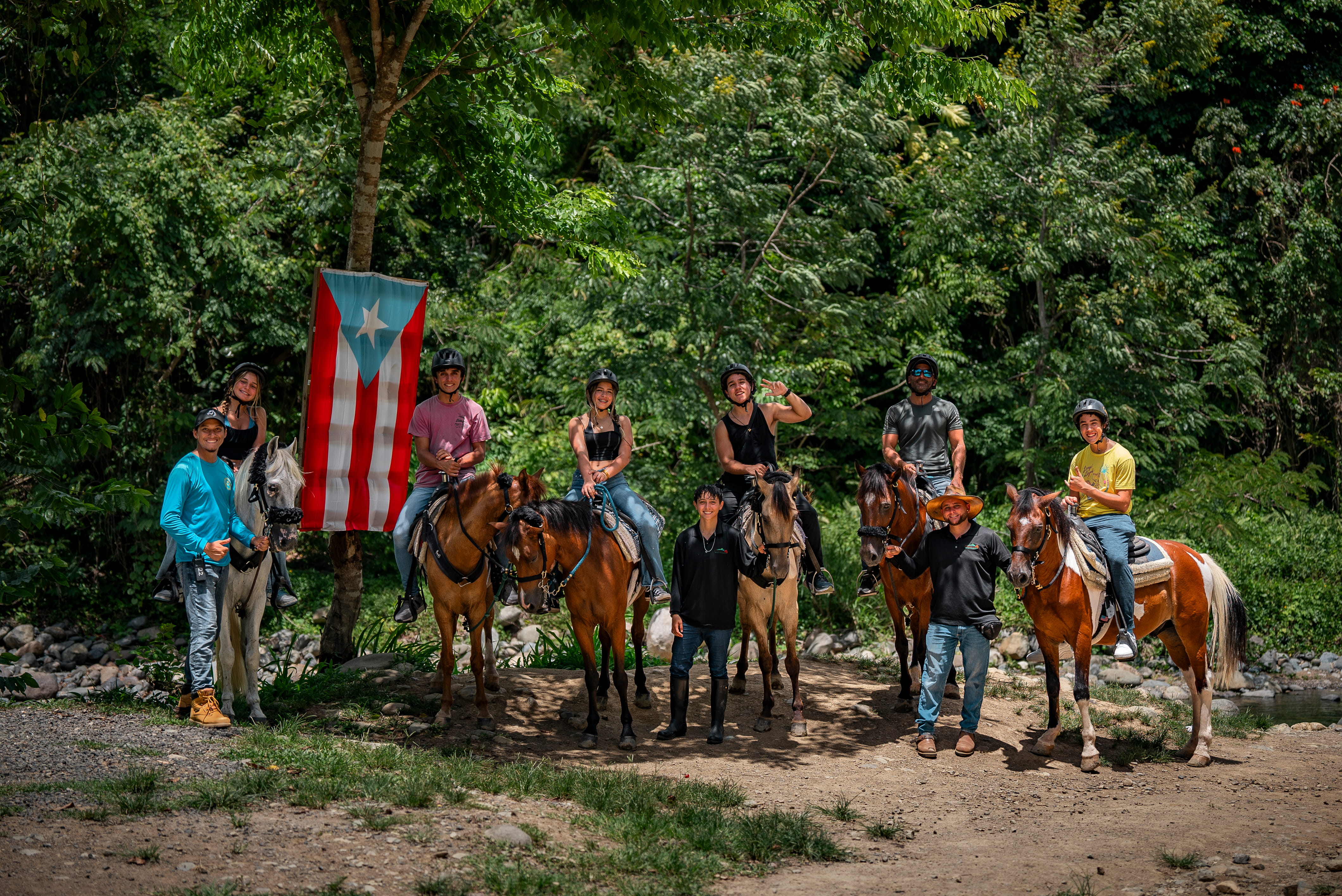 Group of people on horses with a Puerto Rican flag in a forested area.