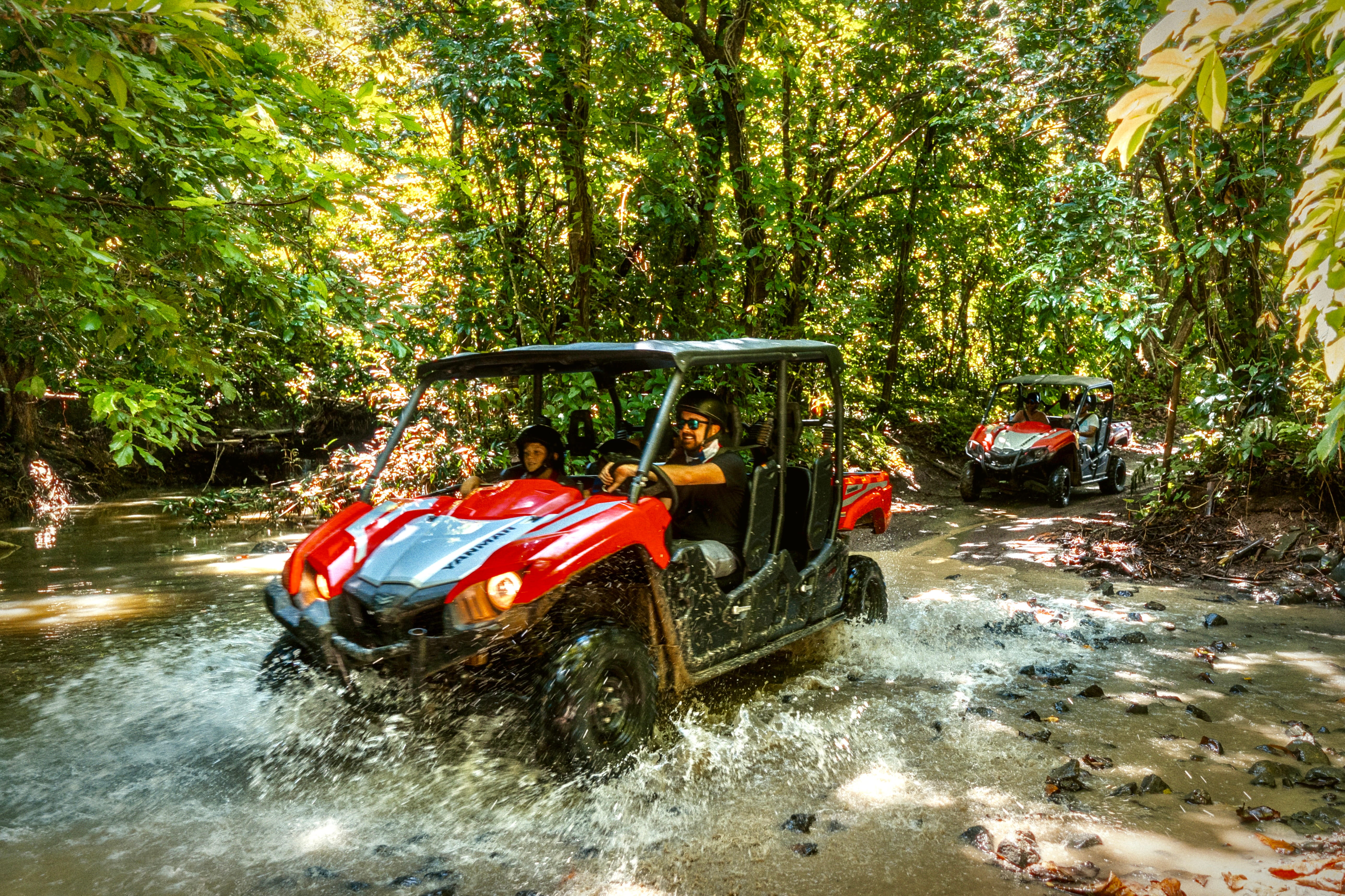 Two red all-terrain vehicles driving through a forest stream.