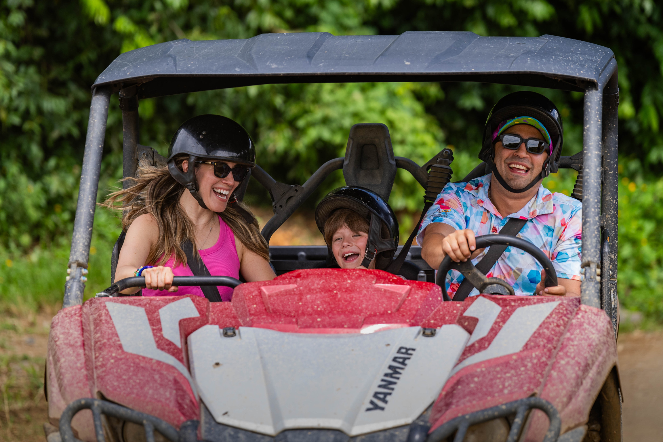 Three people in helmets smiling while riding a red dune buggy.