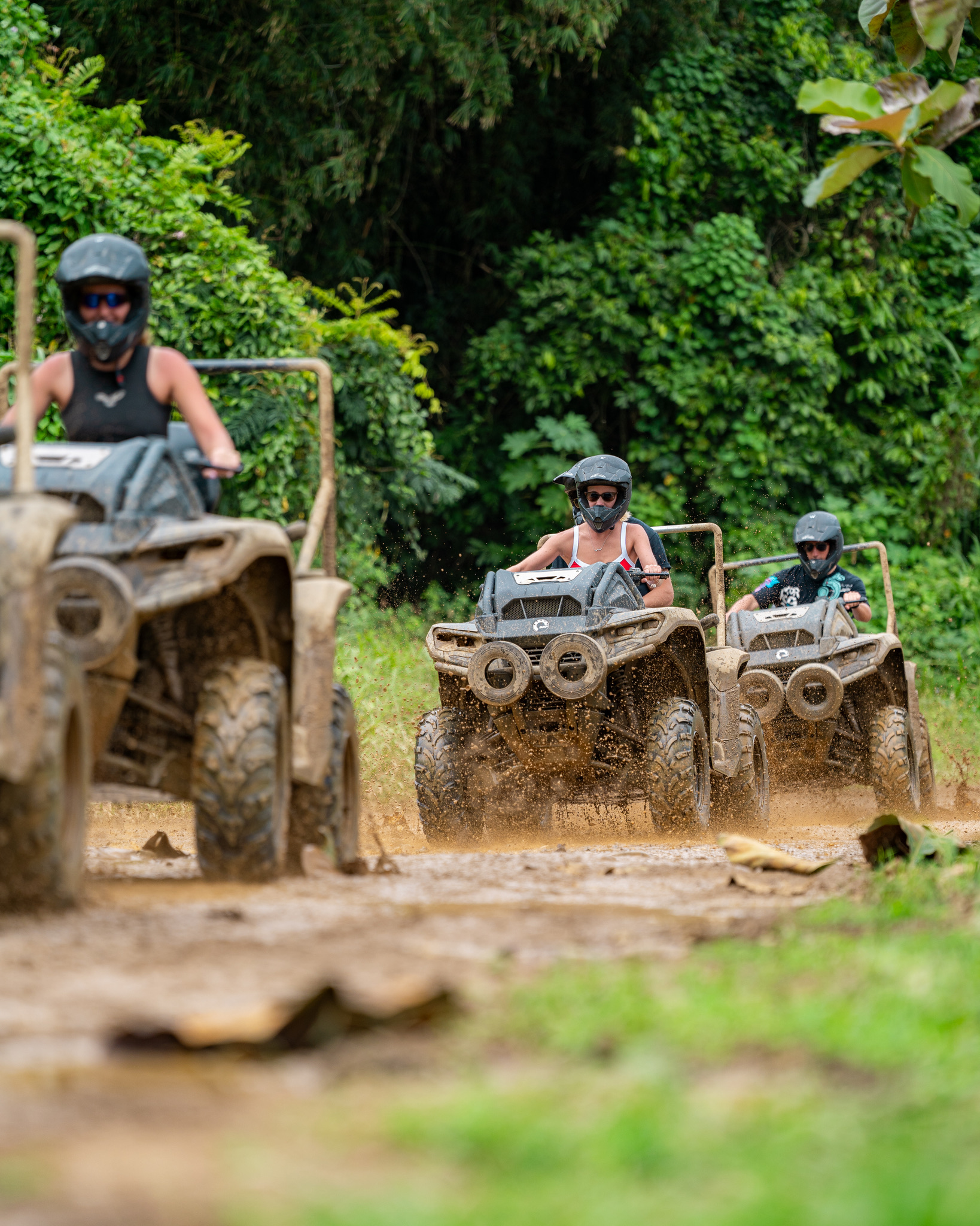 Three people driving off-road vehicles on a muddy trail in a forest.