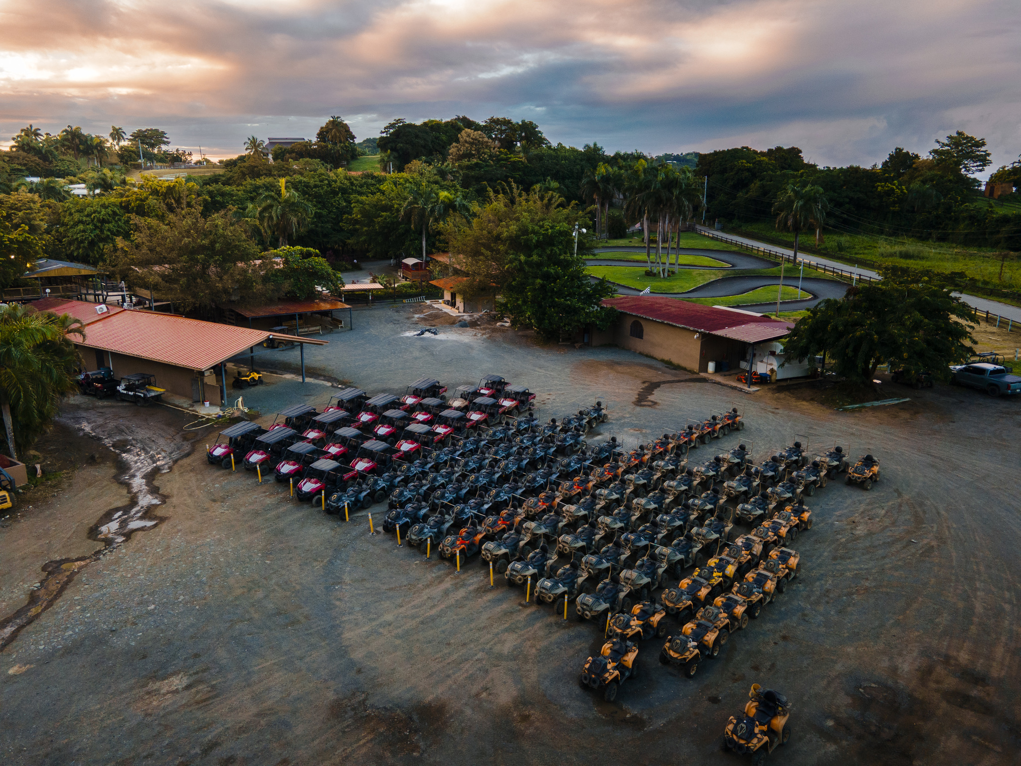 Aerial view of parked ATVs in rows beside buildings and trees under a cloudy sky.