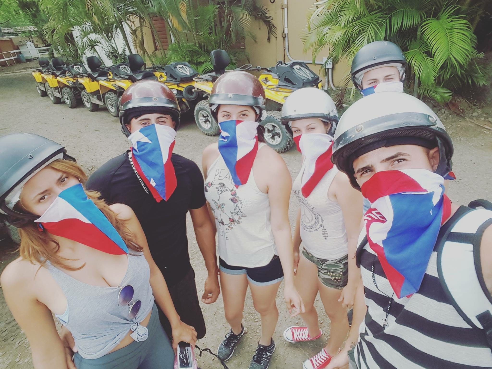 Group of people in helmets and masks with flag design, standing near yellow ATVs.
