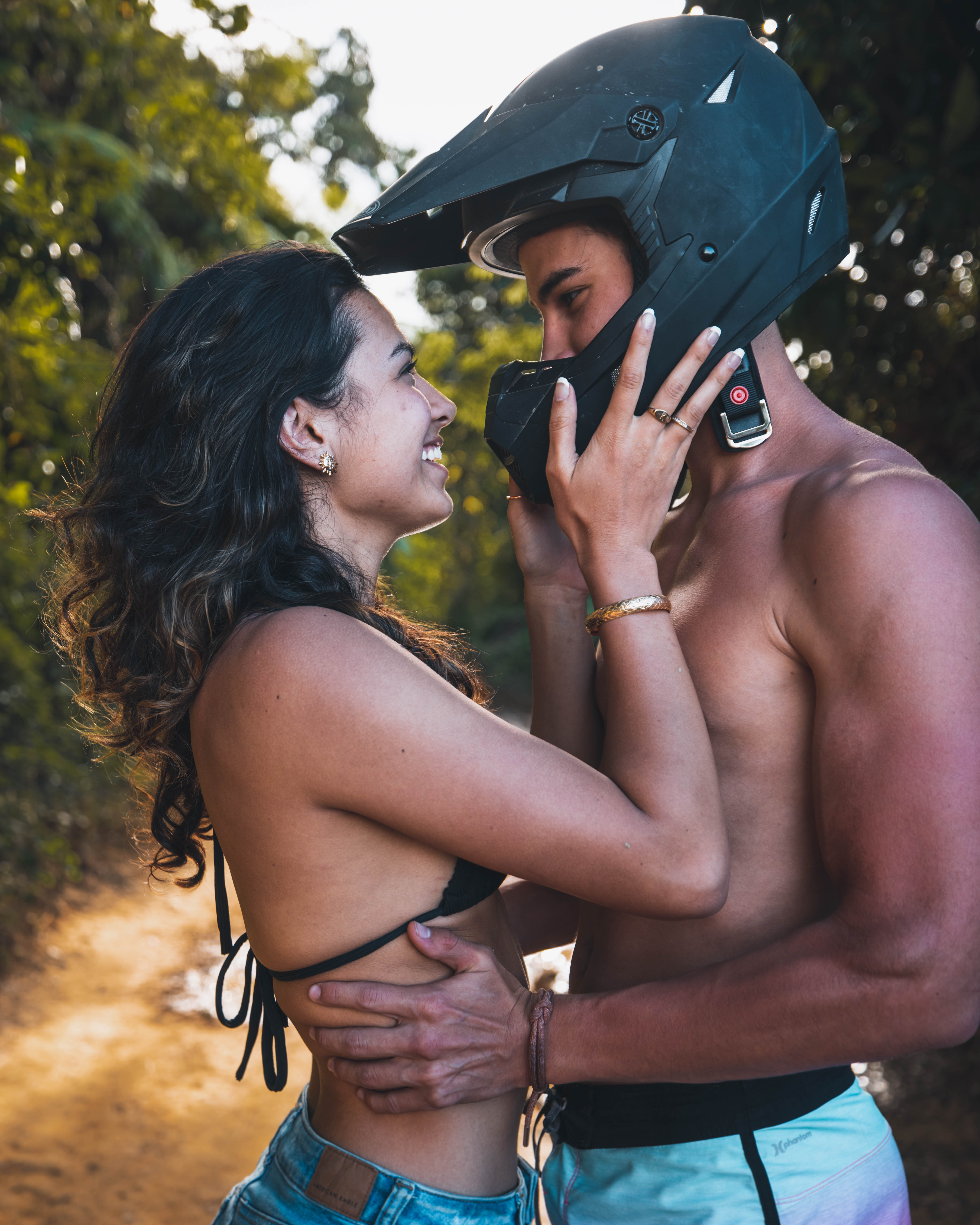 Woman smiling, embraces shirtless man wearing motorcycle helmet on a forest path.