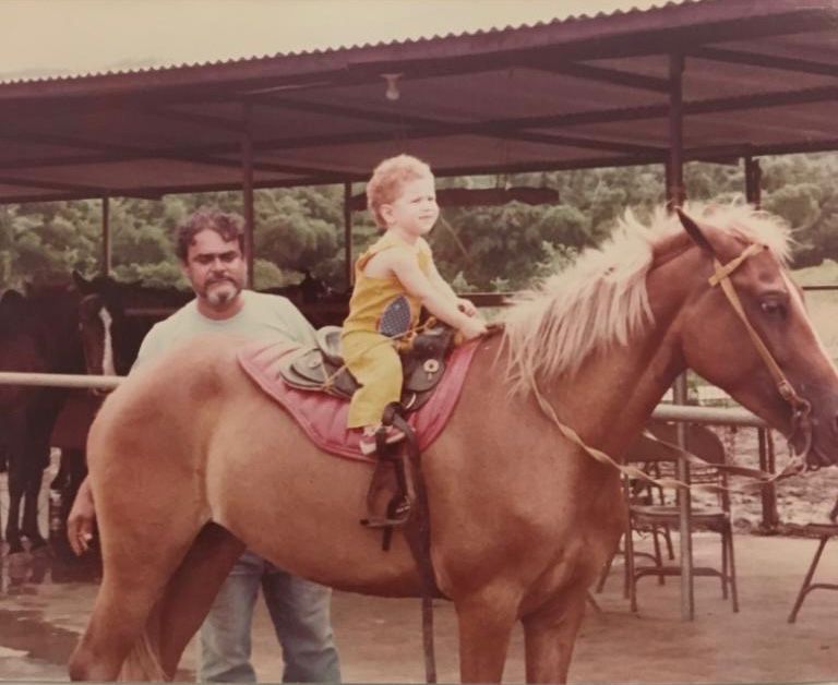 Child in yellow outfit sitting on a horse, guided by a man under a shelter.