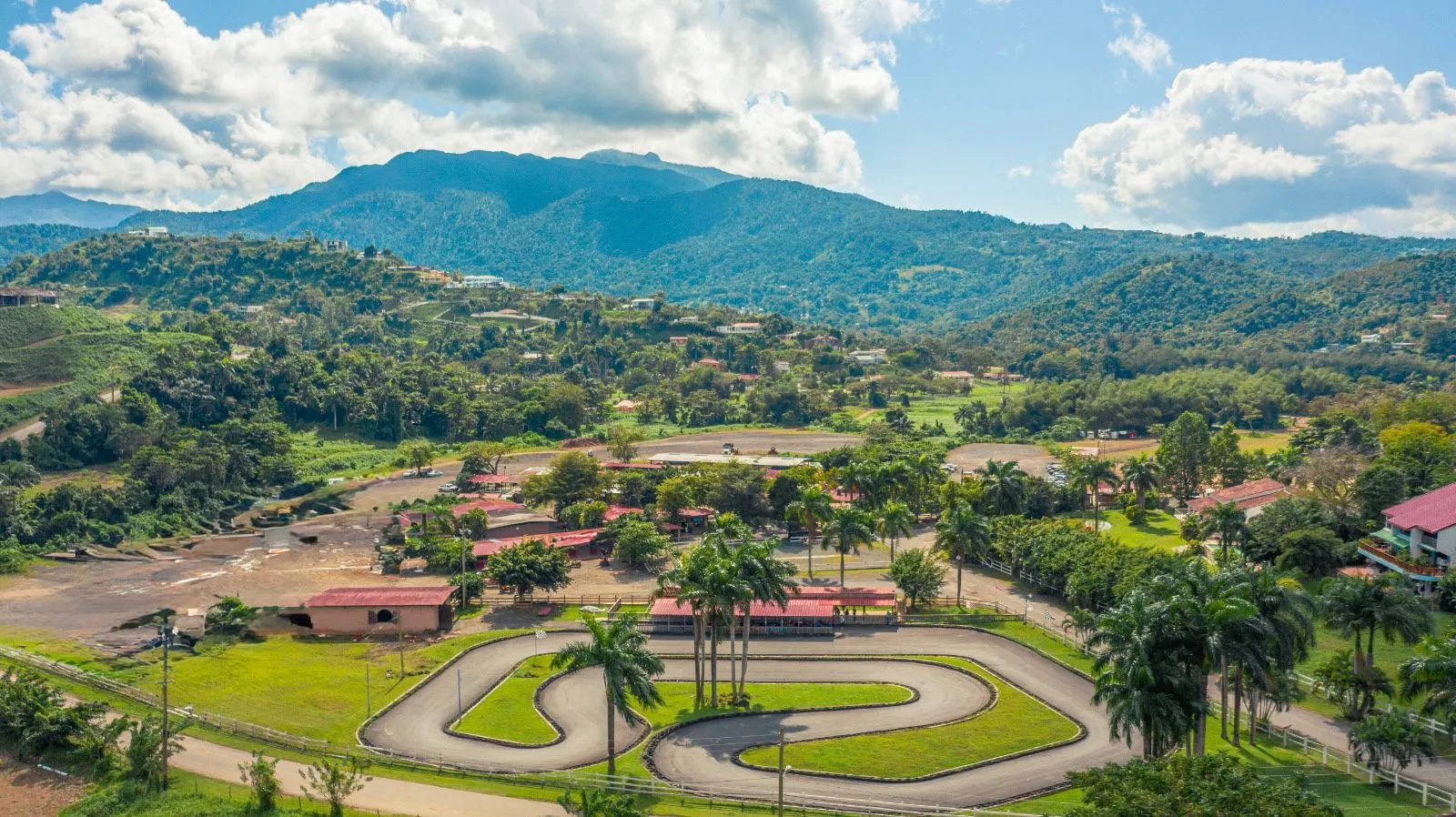 Aerial view of lush landscape with mountains, a go-kart track, and a few red-roofed buildings.