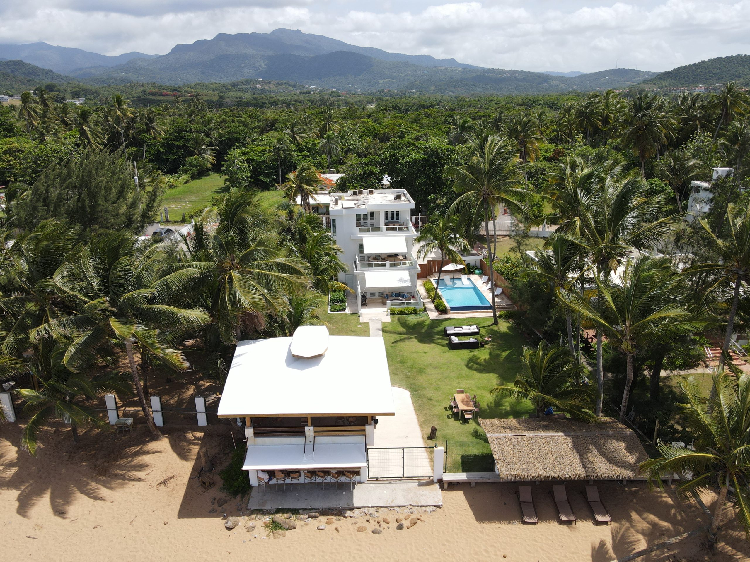 Aerial view of beachfront houses, pool, and lush greenery with mountains in the background.