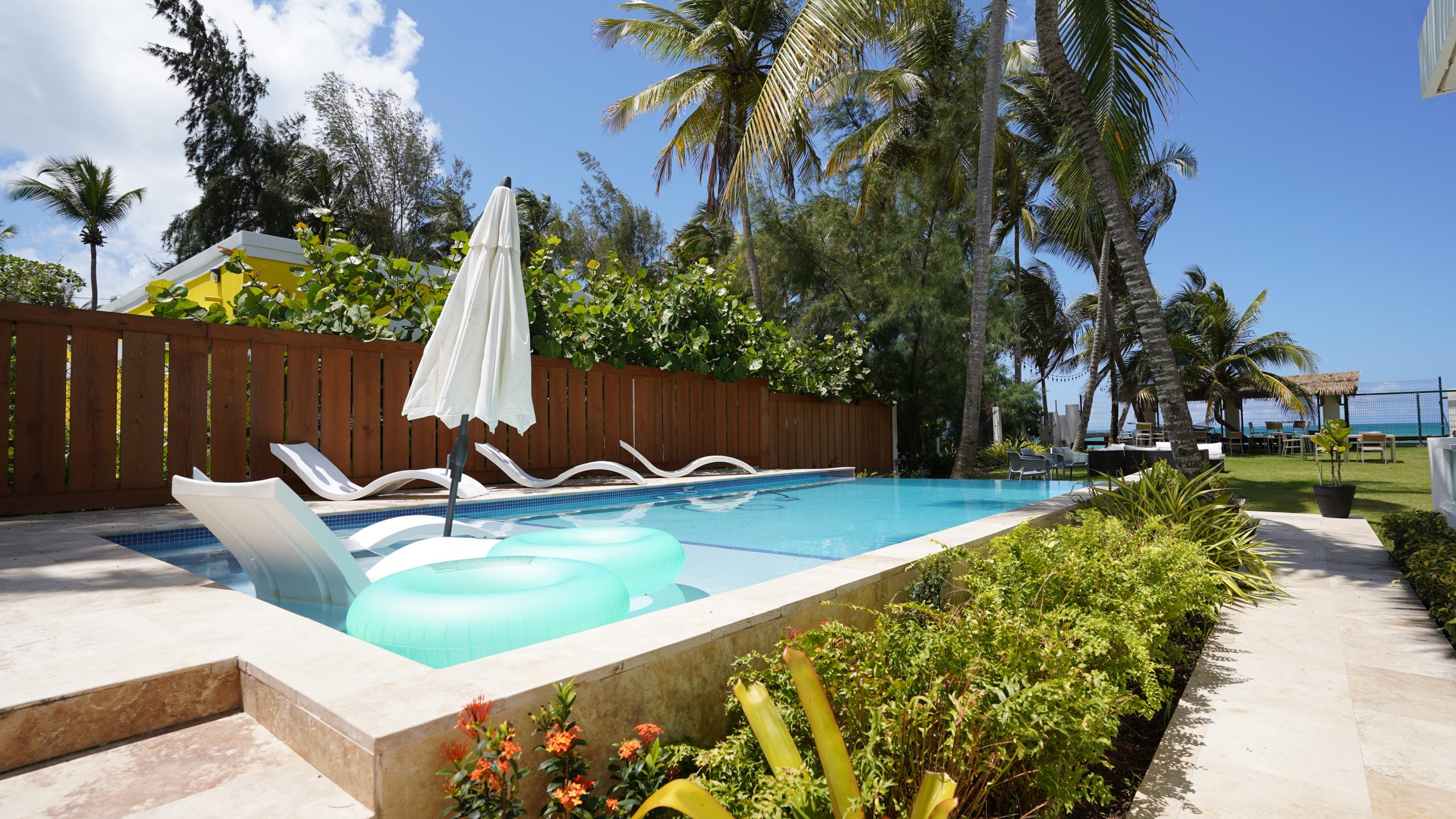 Tropical pool area with loungers, umbrella, and palm trees under a blue sky.