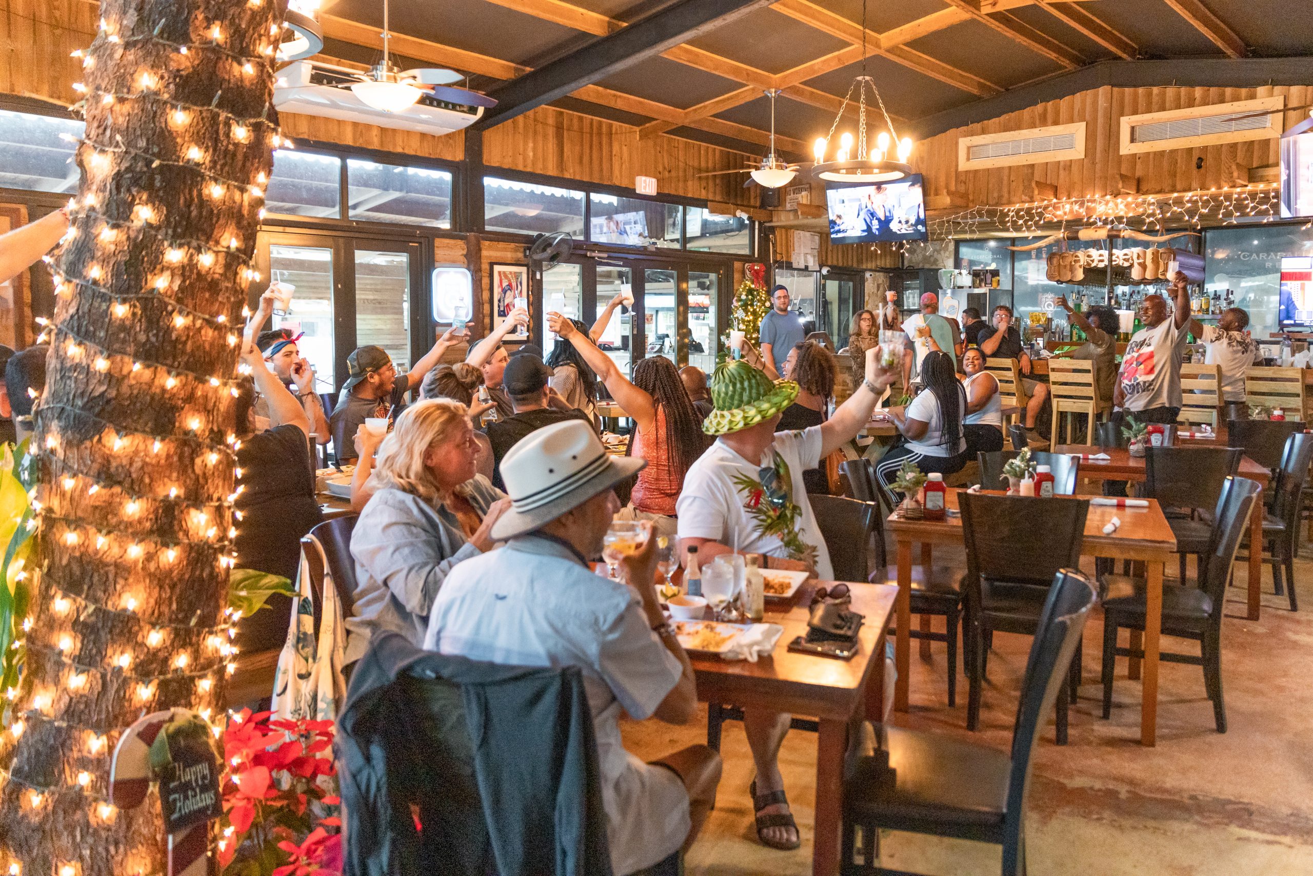 Lively restaurant scene with people eating, tropical decor, and TV screens.