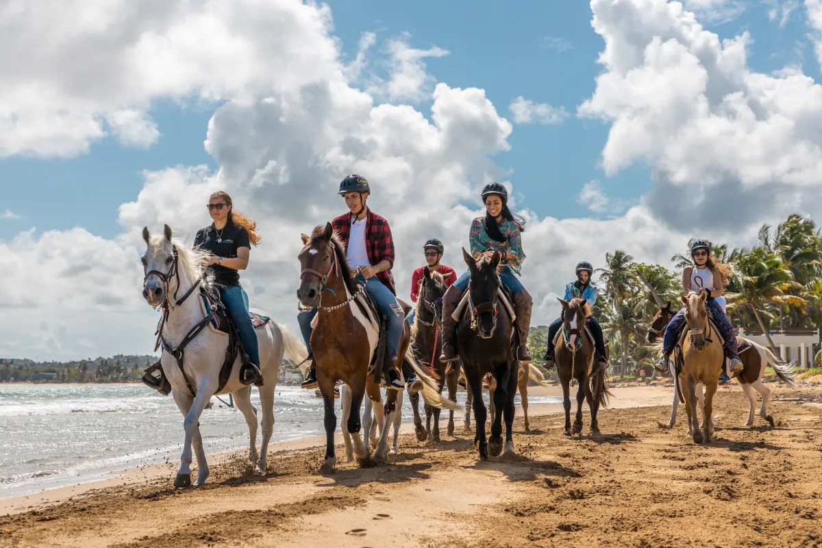 Grupo de personas montando a caballo en una playa de arena bajo un cielo parcialmente nublado.