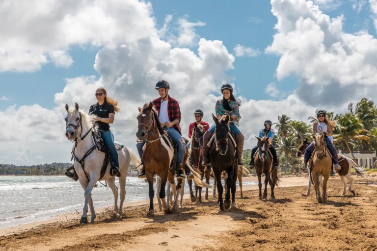 Grupo de personas montando a caballo en una playa de arena bajo un cielo parcialmente nublado.