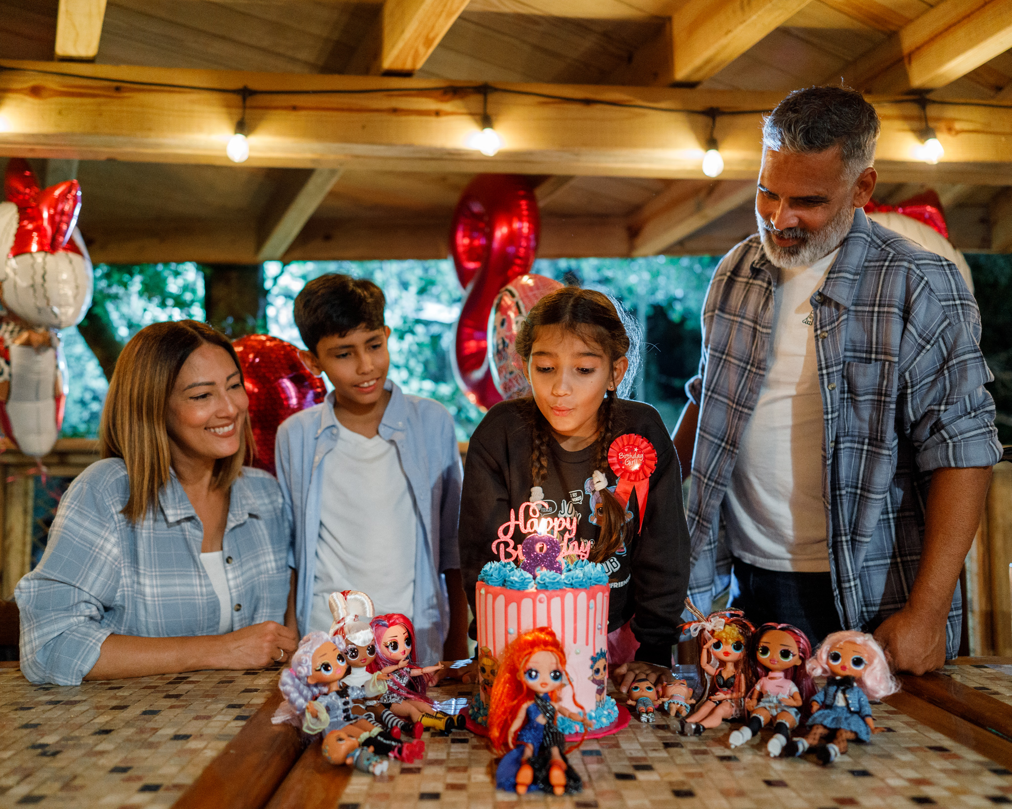 Girl blowing out candles on a birthday cake, surrounded by family and dolls.
